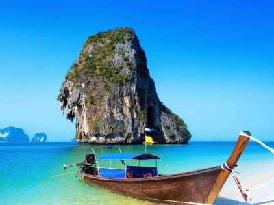 Traditionelles Longtail-Boot an einem weißen Sandstrand in Thailand mit türkisfarbenem Wasser und einer imposanten Felsinsel im Hintergrund.