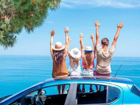 Fröhliche Familie sitzt auf einem blauen Auto und streckt die Arme in die Luft, während sie den Meerblick genießt.