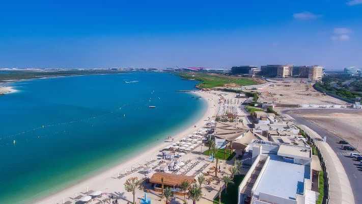 Strandlandschaft mit weißen Sonnenschirmen und Liegen an einem klaren, blauen Meer, im Hintergrund moderne Gebäude und weite Sandflächen, Abu Dhabi