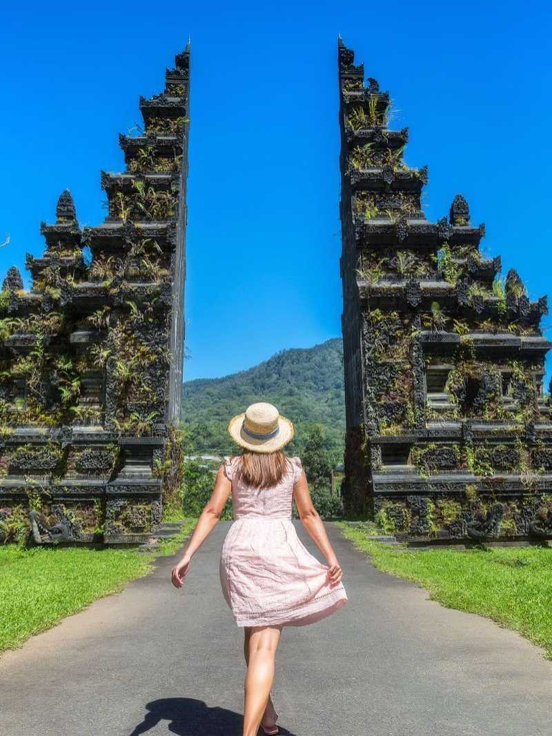 Frau mit Strohhut und Sommerkleid steht vor dem berühmten Handara-Tor auf Bali, umgeben von üppigem Grün und blauem Himmel