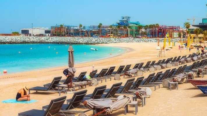 Sonniger Strand in Dubai mit zahlreichen Liegen und Sonnenschirmen, türkisfarbenes Meer und Freizeitpark im Hintergrund, umgeben von Palmen