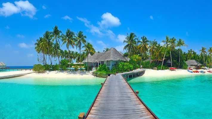 Türkisblaues Wasser mit Holzsteg zu einer tropischen Insel, umgeben von Palmen und Strandbungalows auf den Malediven