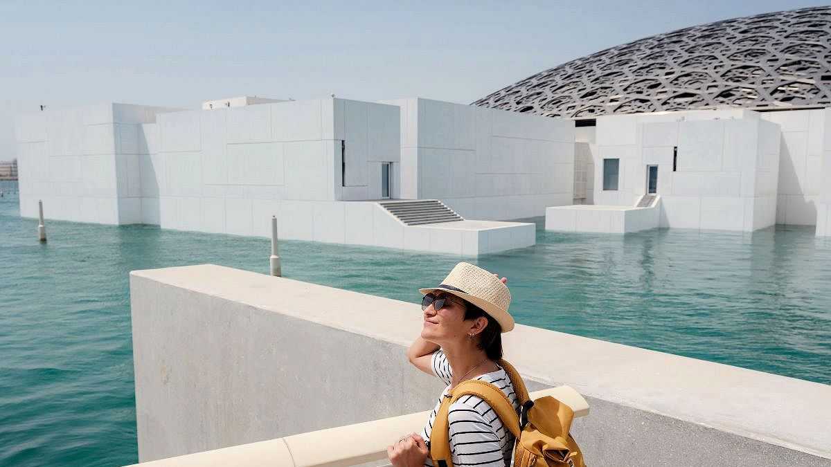 Touristin mit Strohhut und gelbem Rucksack vor dem Louvre Abu Dhabi, umgeben von Wasser und moderner Architektur