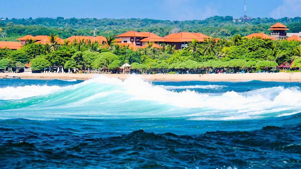 Blick auf den Nusa Dua Beach auf Bali mit Wellen, tropischer Vegetation und Resorts im Hintergrund